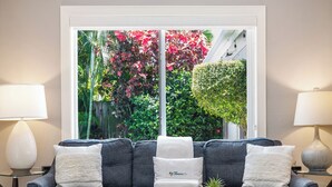 A grey sectional sofa with white and grey cushions, a small round table with a plant, and a window with a view of lush greenery and red foliage. - Modern Tropical Oasis, Private Heated Pool, Near Beaches! (Wilton Manors)