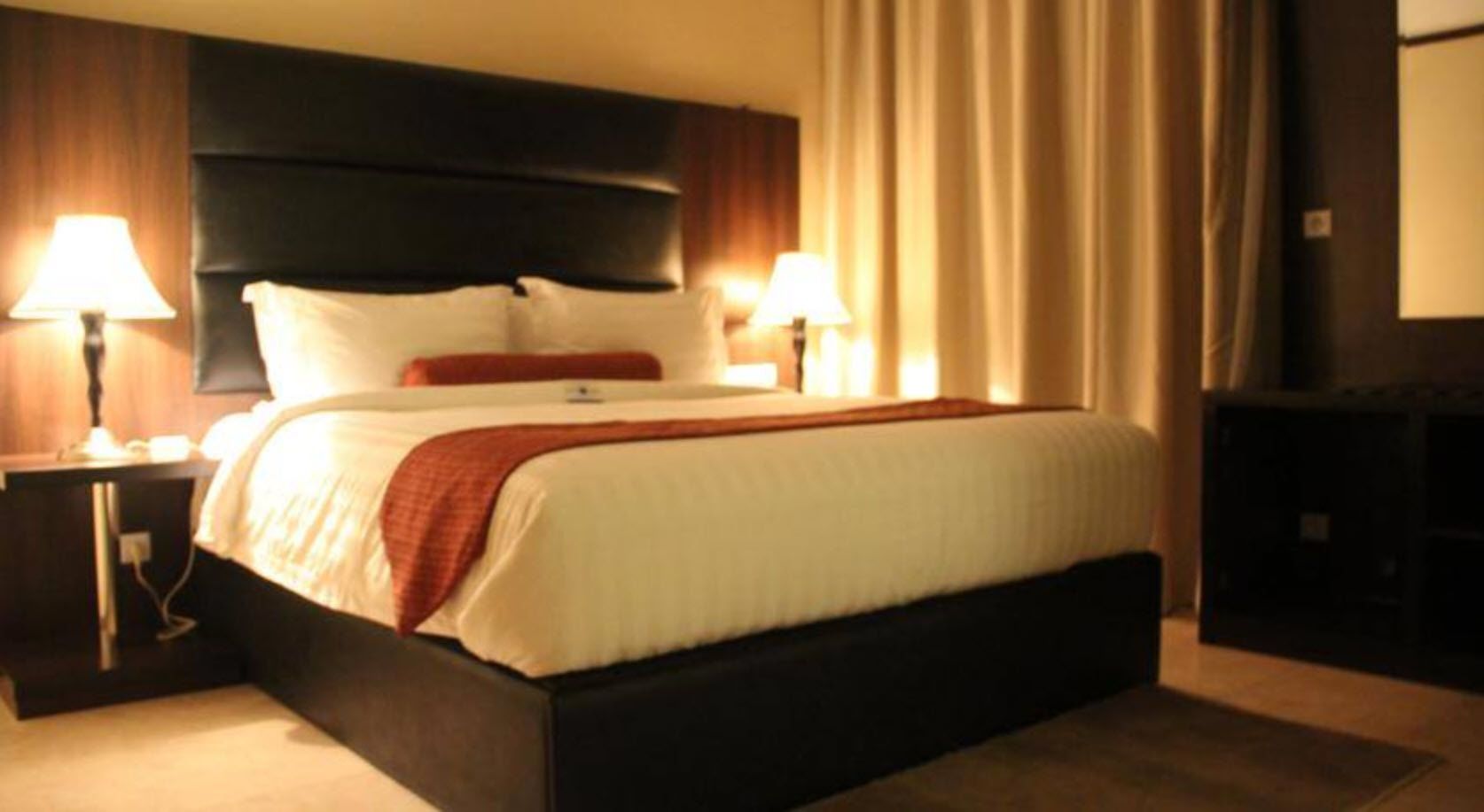 Habitación de hotel con una cama grande, dos mesitas de noche con lámparas, un cabecero oscuro y vistas a un paisaje urbano por la ventana.