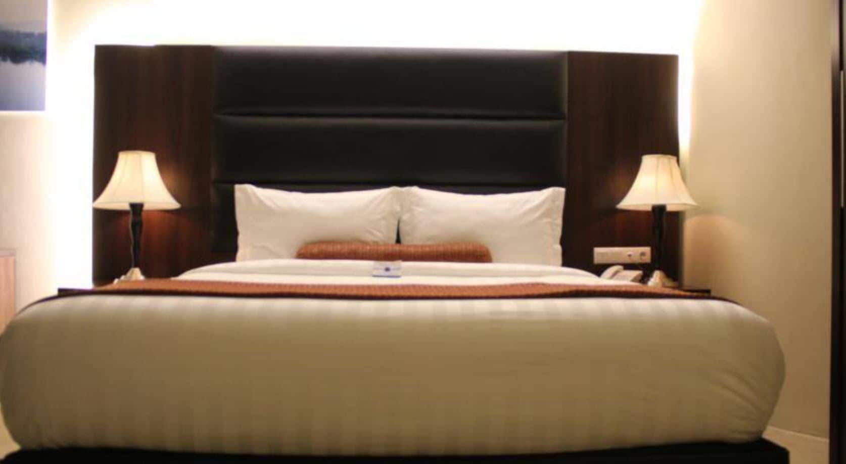 Una cama bien hecha con un cabecero oscuro, dos lámparas de noche y un despertador visible en la mesita.