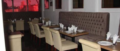 Un restaurante con un largo banco acolchado, mesas individuales con sillas y vistas a palmeras a través de las ventanas.