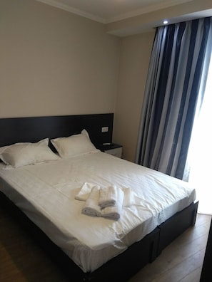 A neatly made bed with white linens and towels, a dark headboard, a nightstand with a lamp, and a window with striped curtains. - Apartment on Marjanishvili Str. (Tbilisi)
