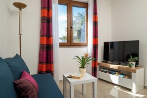 Un salon avec un canapé, une table sur laquelle se trouvent un livre et une plante, un téléviseur posé sur un meuble en bois, et une fenêtre avec des rideaux.
