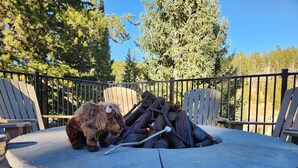 A round stone fire pit with logs and a stick, surrounded by Adirondack chairs on a deck with a metal railing. - Upscale Indian Themed PENTHOUSE On Ski Access Level! Vaulted ceiling. High rated (Winter Park)