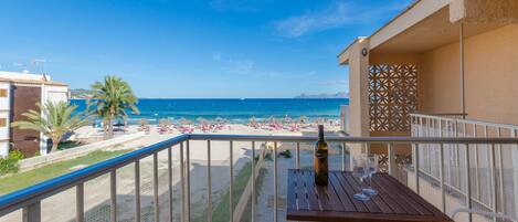 A balcony with a table and chairs overlooking a beach with umbrellas and people.