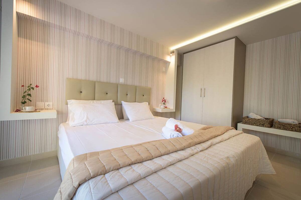 Une chambre d’hôtel moderne avec un grand lit, une tête de lit dotée d’un panneau de couleur claire, une armoire et une petite étagère sur laquelle est posé un vase de fleurs.