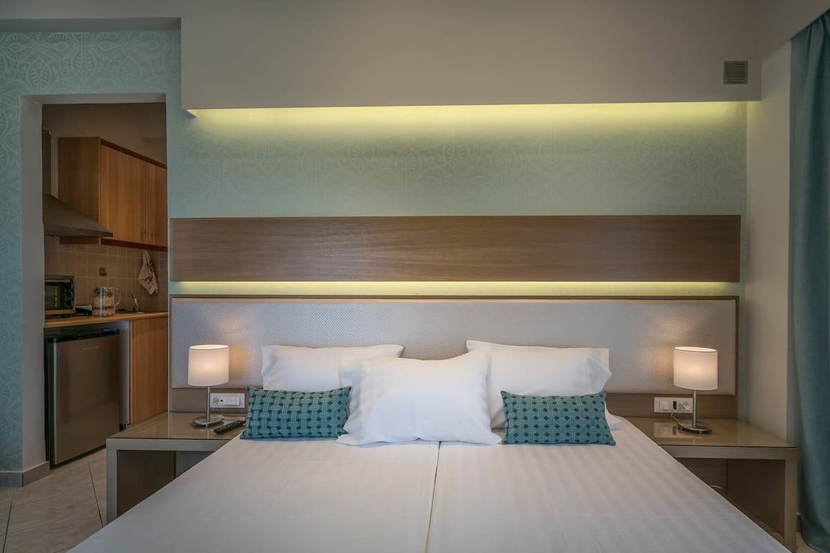 Une chambre d’hôtel avec un grand lit, deux tables de chevet avec des lampes, une tête de lit en bois, une kitchenette et un mur à motifs.