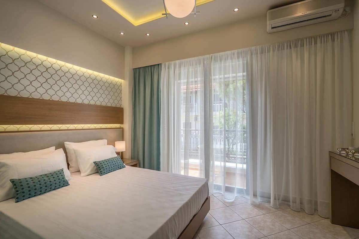 Une chambre d’hôtel avec un grand lit, une table de chevet avec une lampe, un luminaire au plafond, un appareil de climatisation et une vue sur l’extérieur à travers des rideaux légers.