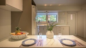 A modern kitchen with a dining table set for two, featuring a fruit basket and a vase with greenery. - Alba Boutique Apartments (Zakynthos)