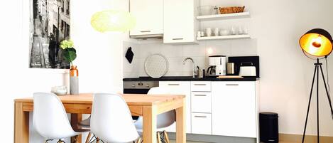 Een moderne keuken met witte kasten, een houten eettafel met vier stoelen, een zwarte prullenbak en een vloerlamp.