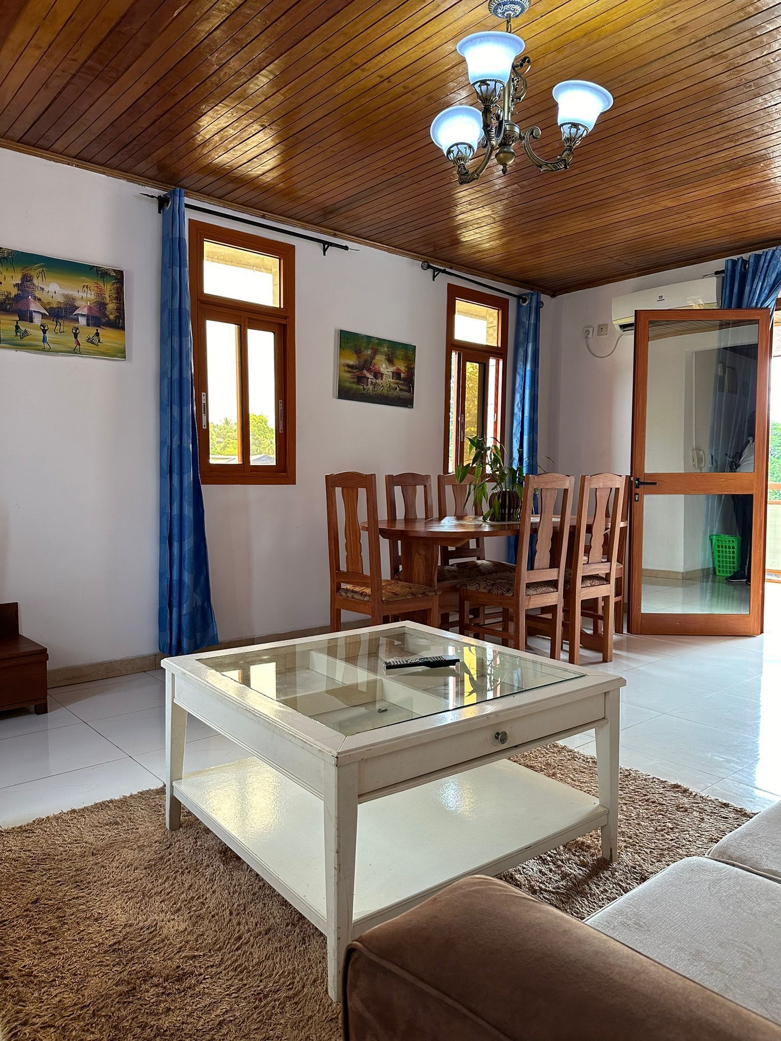 A room with a wooden ceiling, a glass-top table, wooden chairs, a brown sofa, and two windows with blue curtains.