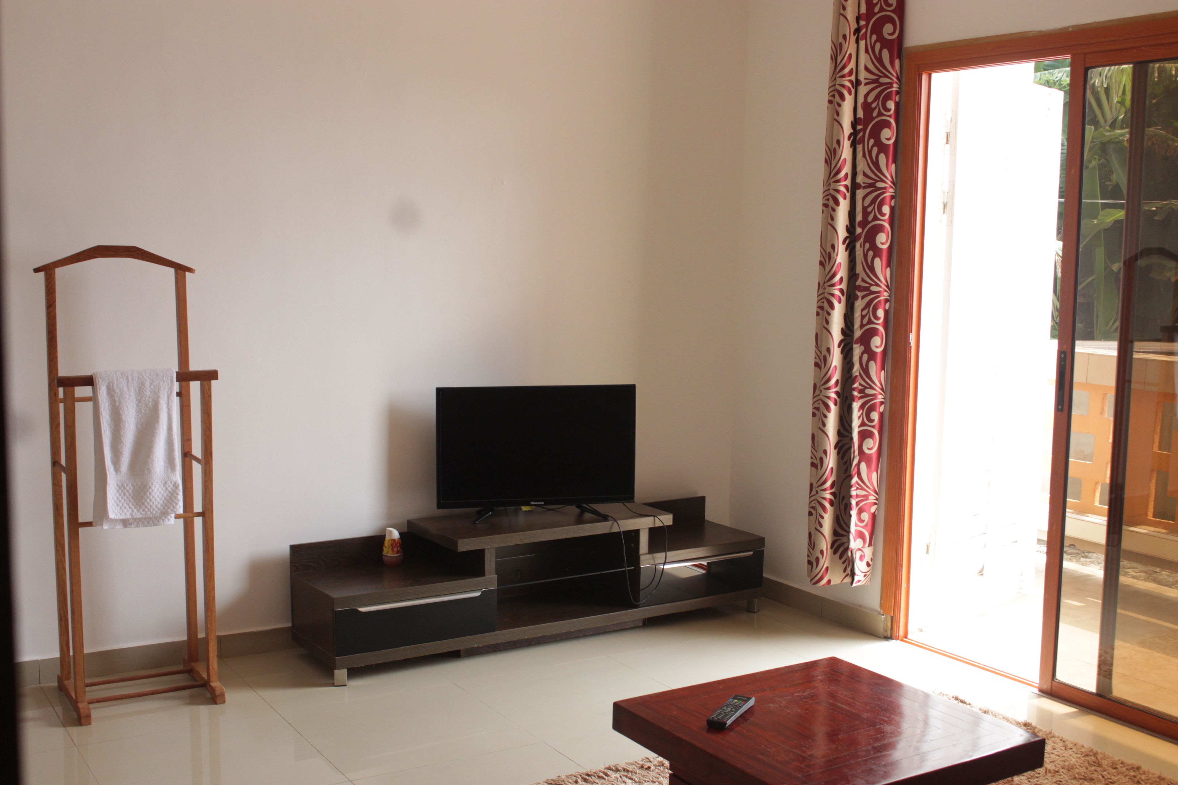 A living room with a flat-screen TV on a wooden stand, a wooden chair with a towel, and a sliding glass door to the outside.