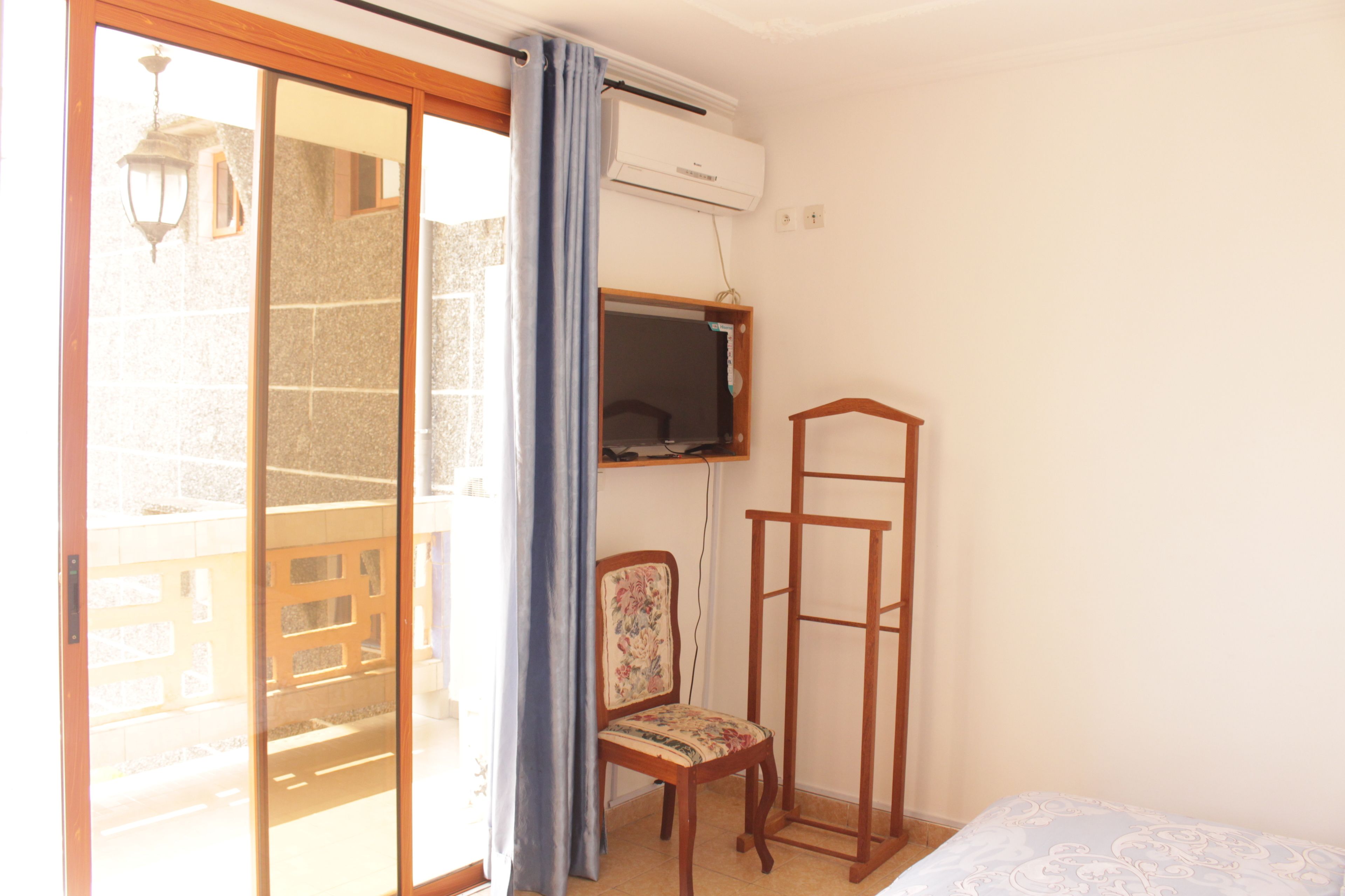 A room with a sliding glass door leading to a balcony, a wooden chair, a TV mounted on the wall, and a bed with a patterned bedspread.