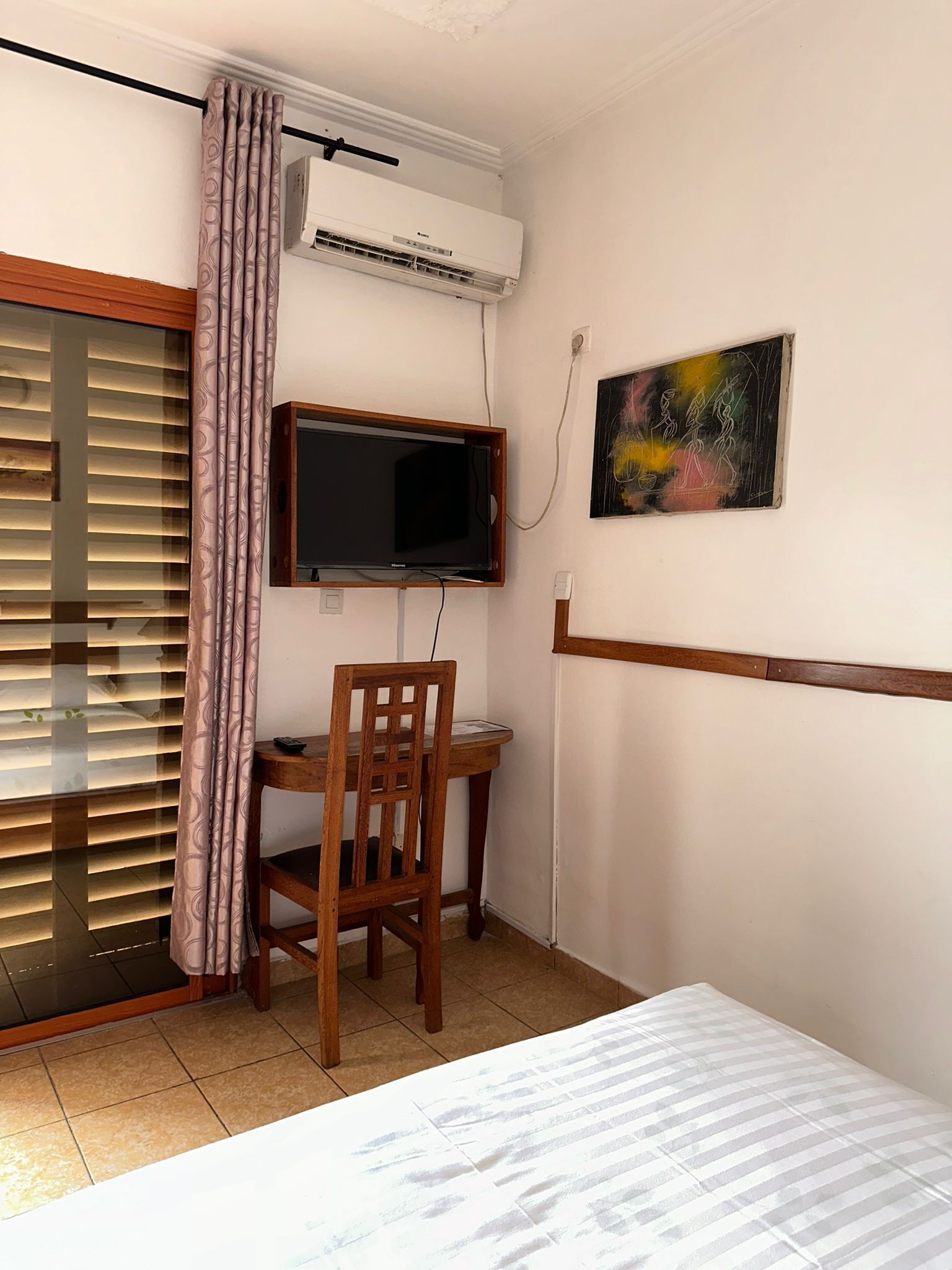 Una habitación con cama, escritorio y silla, aire acondicionado, televisor y un cuadro en la pared.