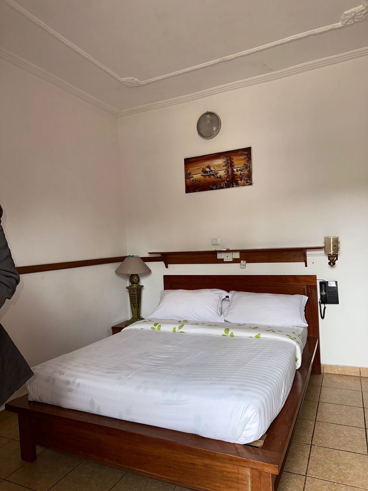 Un lit bien fait, avec du linge de lit blanc orné d’un motif de feuilles vertes, une tête de lit en bois, une lampe de chevet et un téléphone fixé au mur.