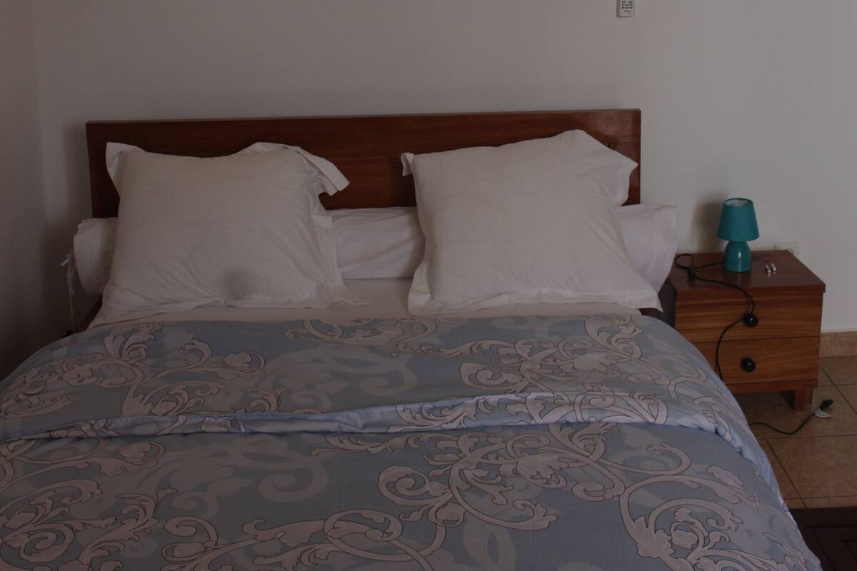 Ein Schlafzimmer mit einem hölzernen Kopfteil, einer gemusterten Tagesdecke, zwei weißen Kissen, einem Nachttisch mit einer Lampe und einer Wandleuchte.