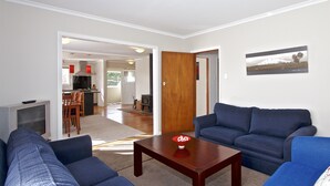 Una sala de estar con un sofá, una mesa de centro y un televisor. Hay una puerta que da a una cocina y un comedor.