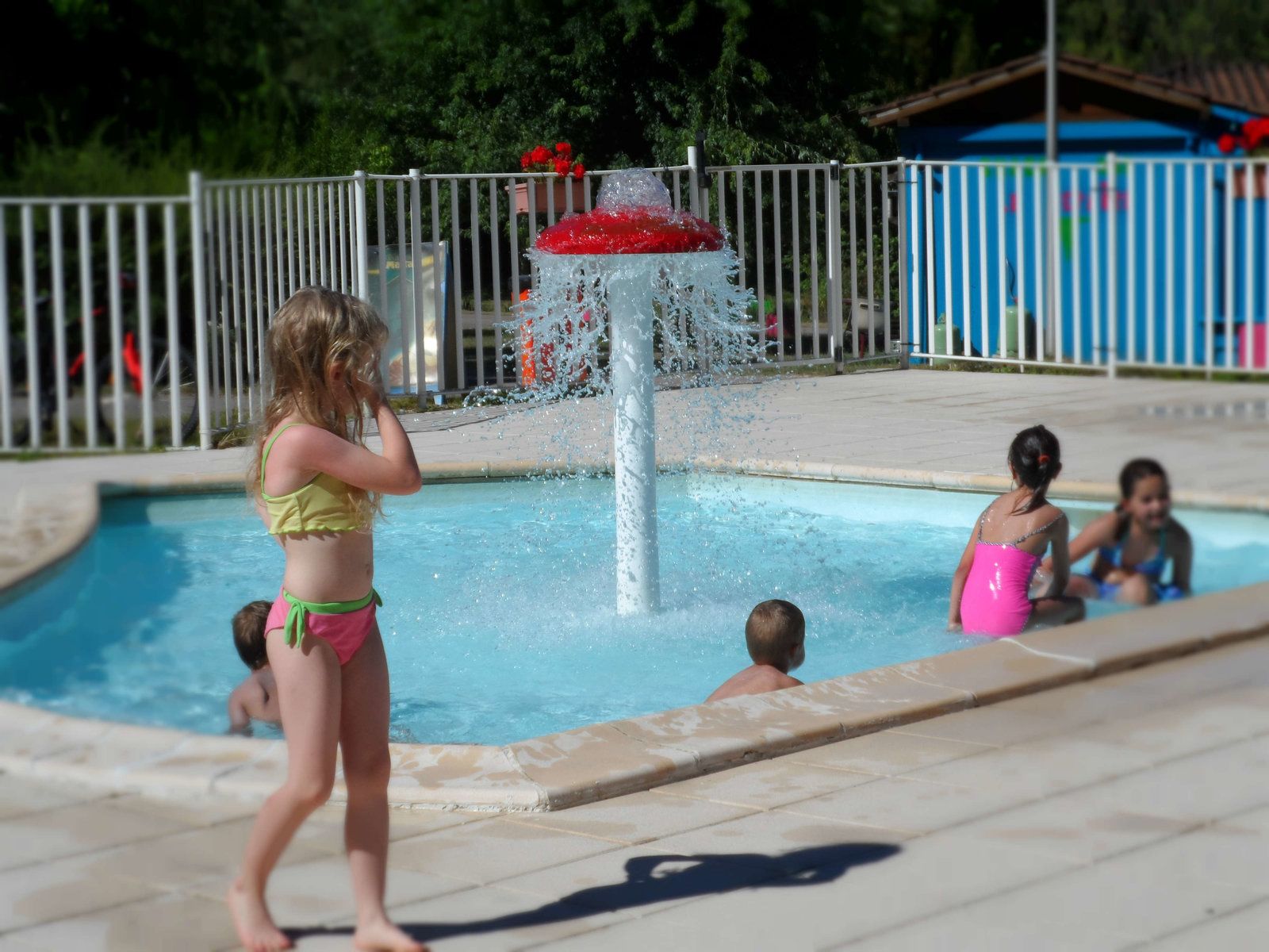A swimming pool area with a water fountain, a fenced playground, and a building in the background.
