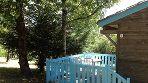 A wooden cabin with a blue deck and white chairs, surrounded by trees. - Camping le Repaire - Les Chalets (Thiviers)