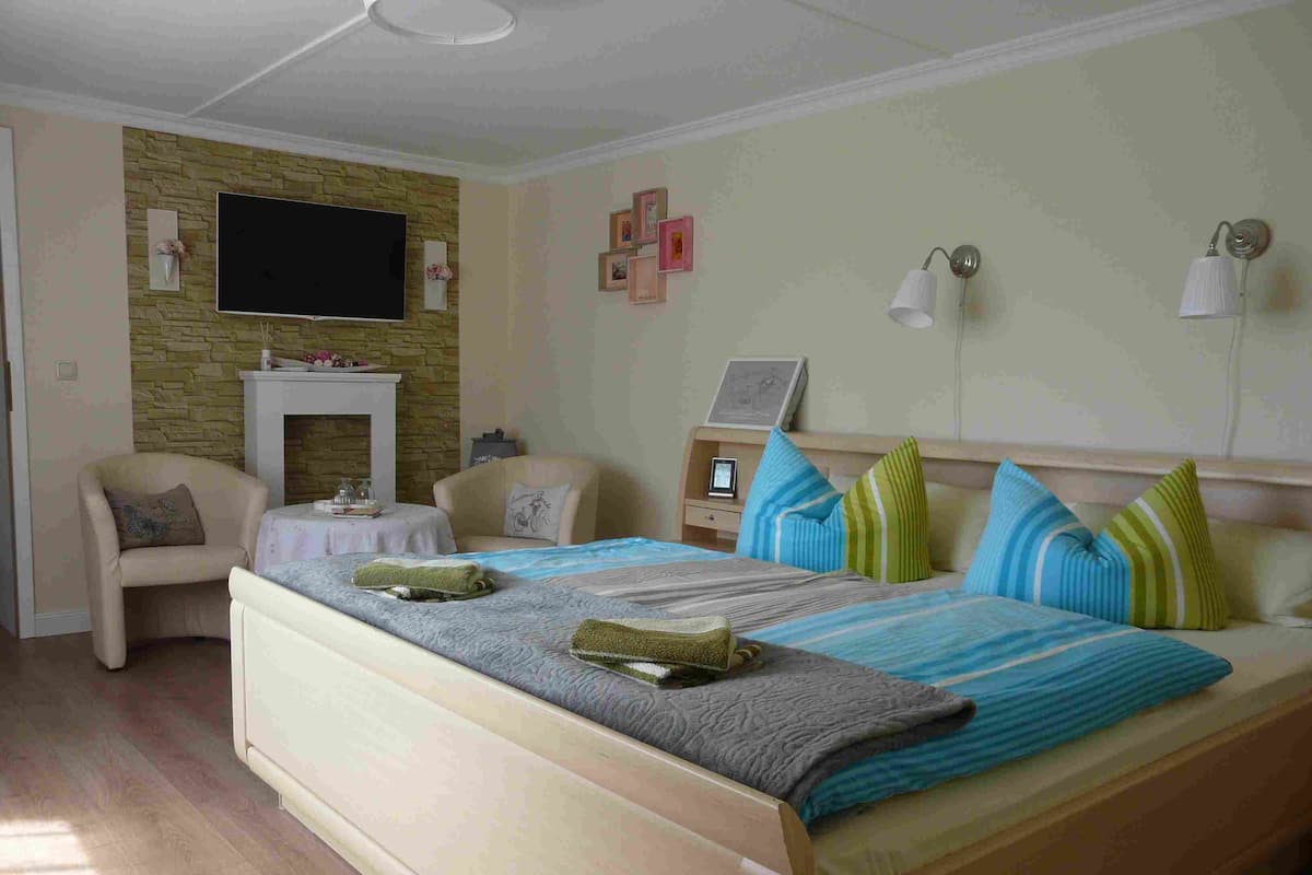 A bedroom with a bed, two chairs, a small table, a fireplace, and a wall-mounted TV.