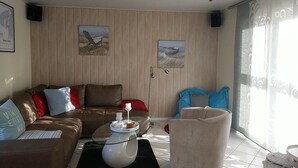 A living room with a brown leather sofa, a glass coffee table, and a beige armchair. There are two framed pictures on the wall, a window with curtains, and a tiled floor. - Pension Zur Kirchsee (Insel Poel)