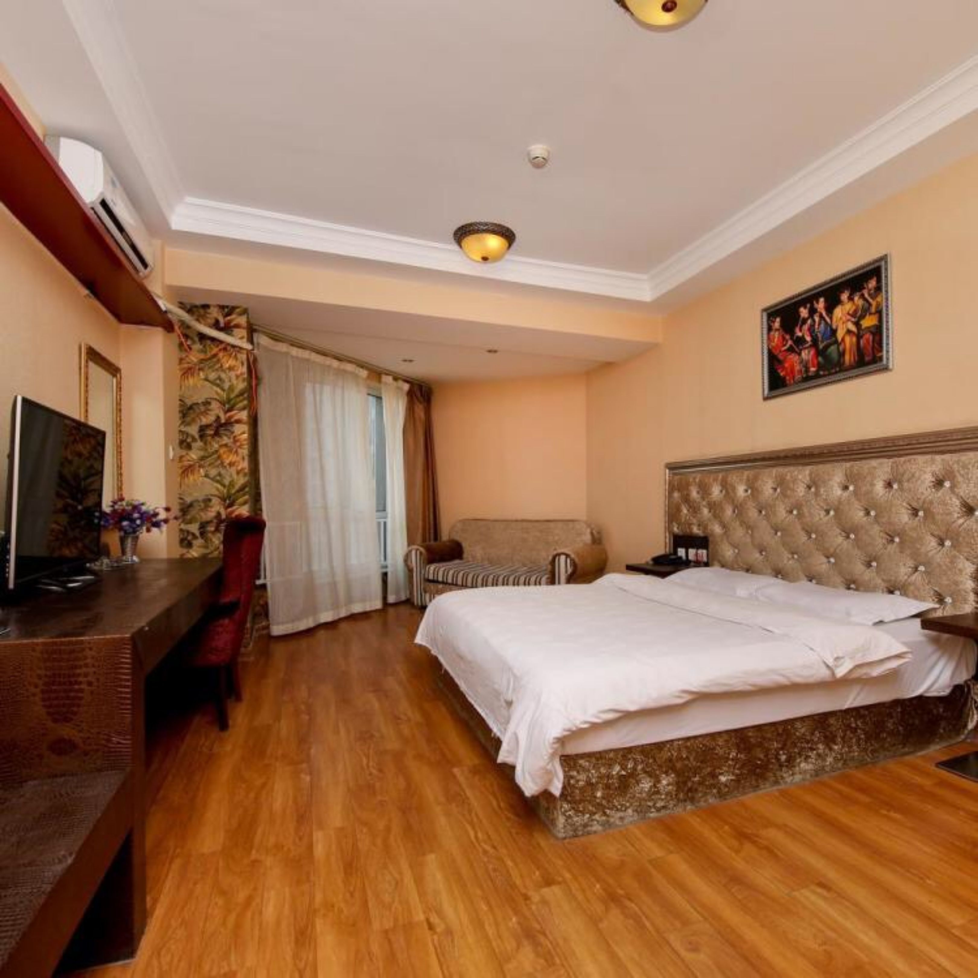 A hotel room with a wooden floor, a bed with a tufted headboard, a desk with a computer, a chair, and a painting on the wall.