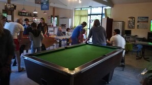 A billiards room with a pool table, people playing, and a bar area in the background.
