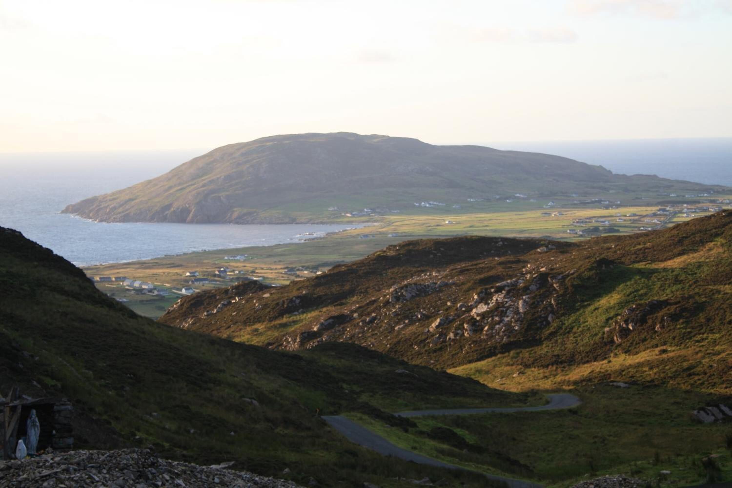 A coastal landscape with a prominent hill, a winding road, and scattered buildings.