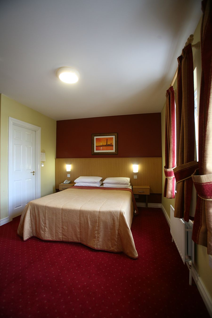 A hotel room with a bed, bedside tables, a nightstand, a painting on the wall, and a window with curtains.
