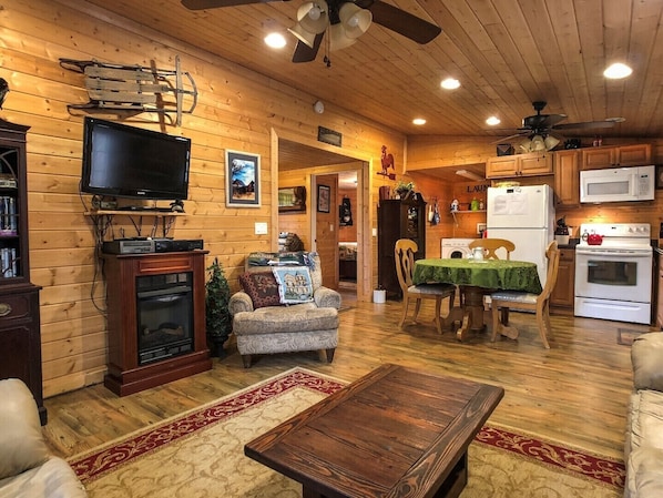 Interior acolhedor de uma cabana de toras, com televisão, lareira, sofá, mesa de jantar e cozinha.