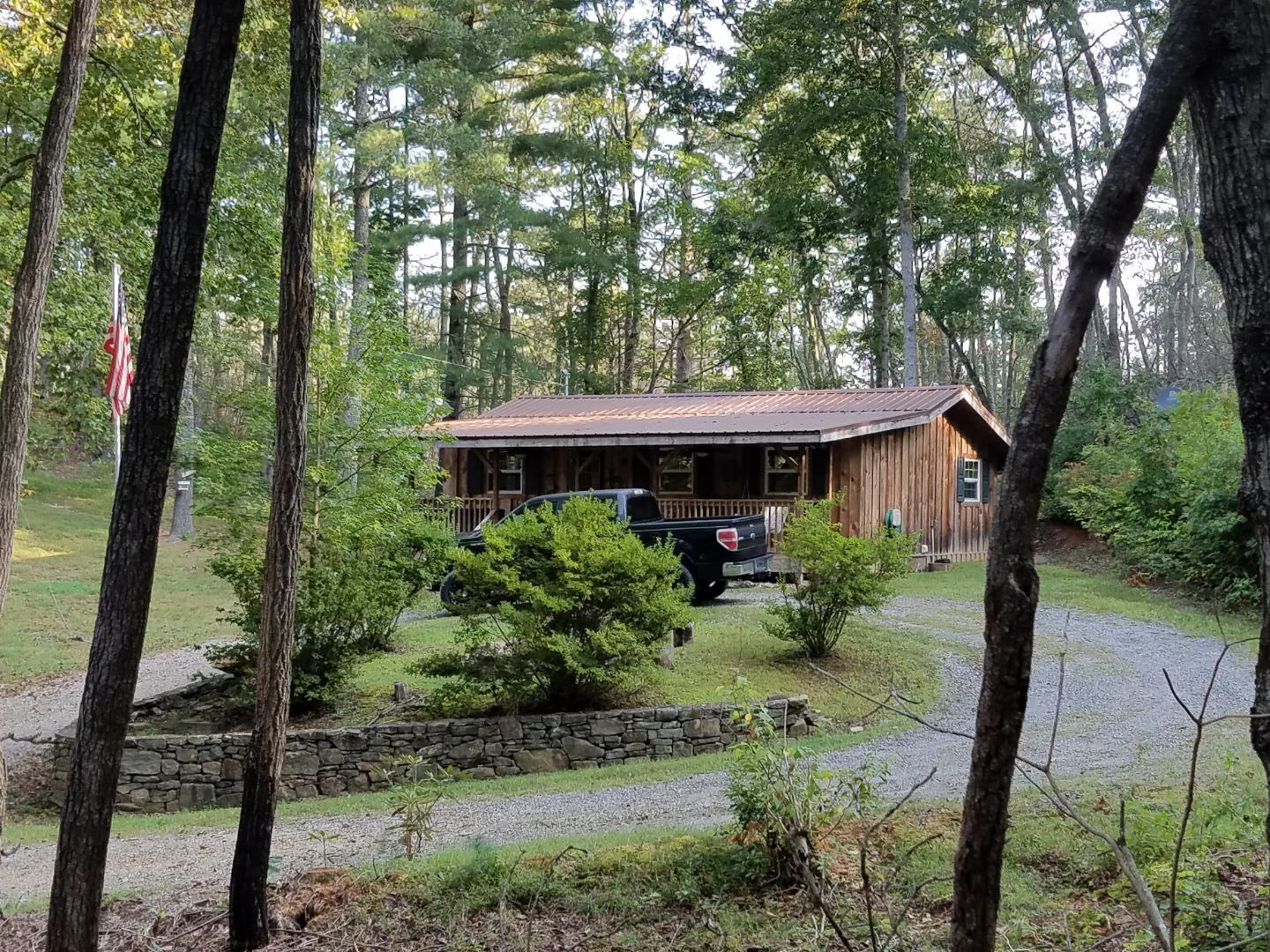 A rustic cabin with a porch, surrounded by trees and a stone wall.