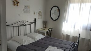 Una cama bien hecha con una colcha oscura y almohadas blancas, una mesita de noche con una lámpara, un espejo en la pared y una ventana con cortinas translúcidas.