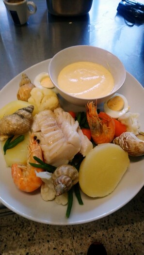 Une assiette de fruits de mer comprenant des crevettes, du poisson et des coquilles Saint-Jacques, accompagnée d’œufs durs, de pommes de terre et d’un bol de sauce.