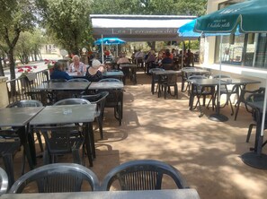 Un café en plein air, avec des tables et des chaises, protégé du soleil par des parasols, et une enseigne indiquant le nom et l’histoire de l’établissement.