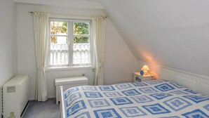 Quarto pequeno e arrumado, com uma cama, janela com cortinas, mesa de cabeceira com abajur e radiador.