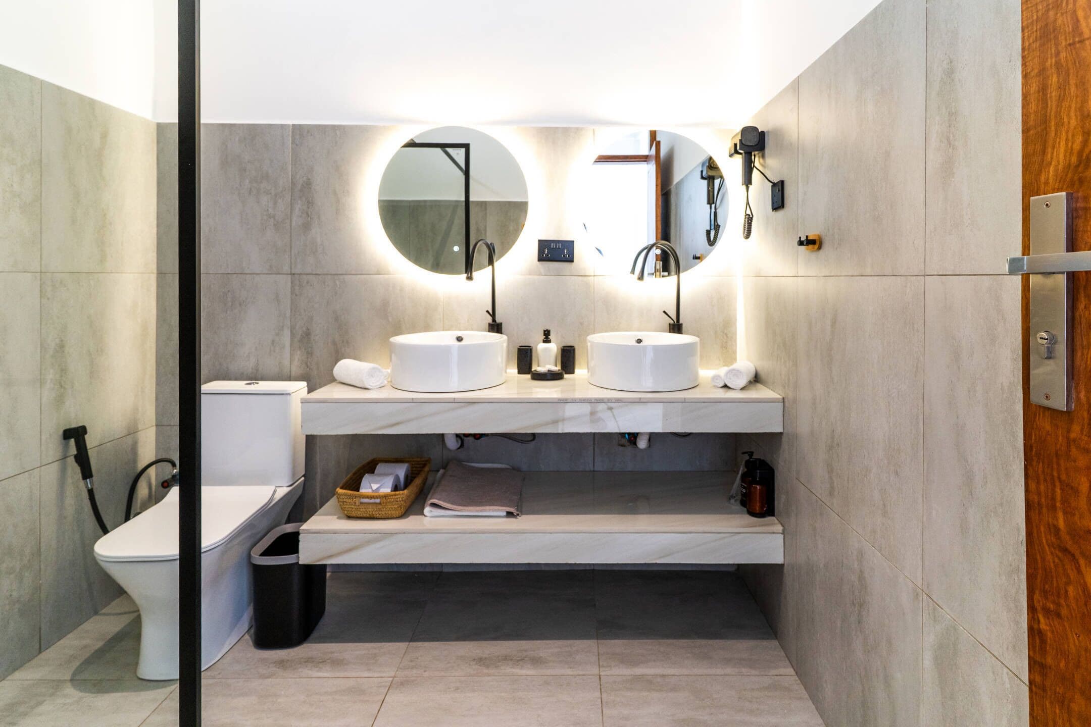 A modern bathroom with two sinks, a toilet, and a bidet. There are mirrors above the sinks, and a towel rack is visible.