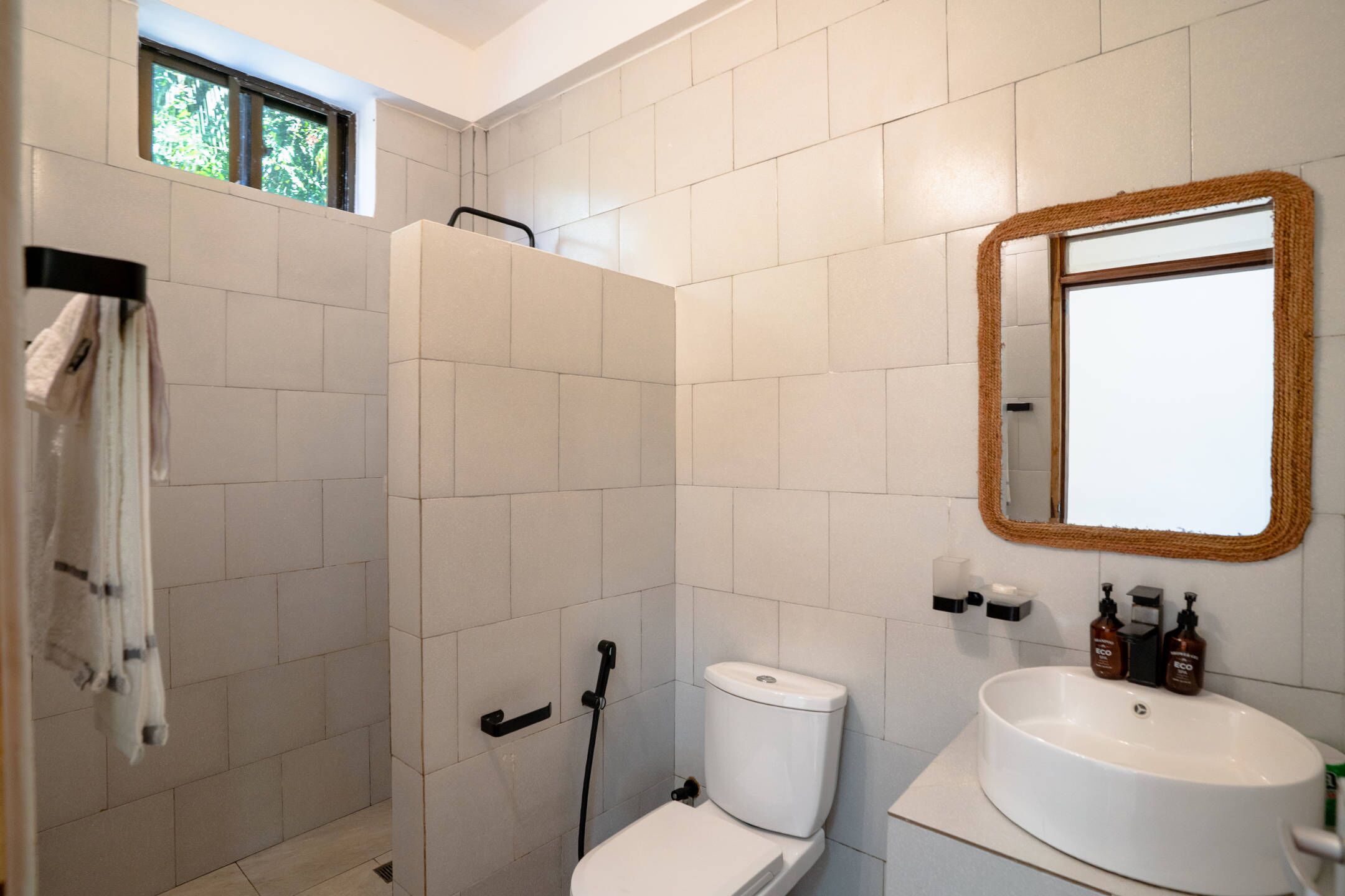 Une salle de bain avec des carreaux blancs, un lavabo blanc, des toilettes, une douche et une fenêtre.