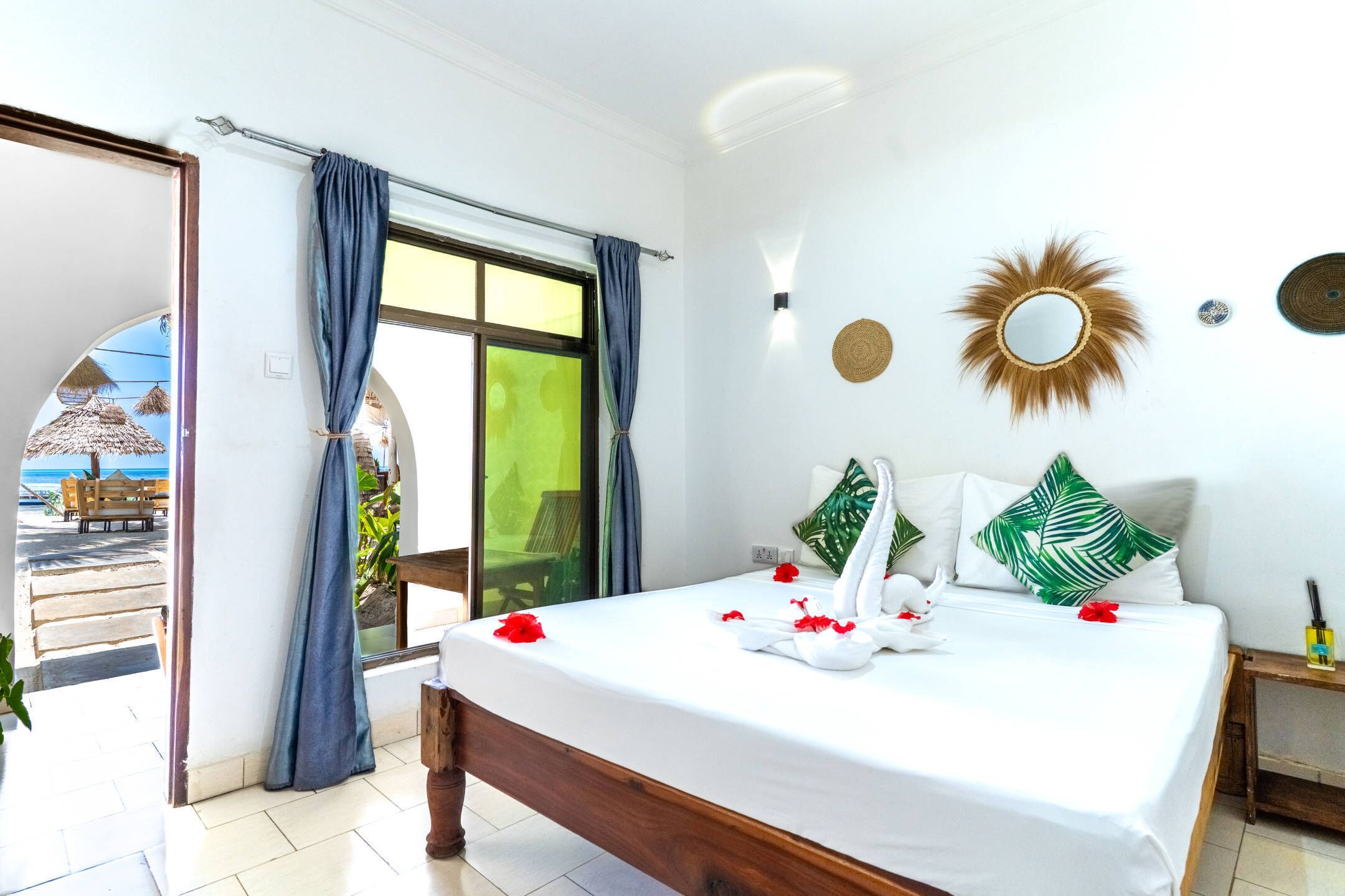 A bedroom with a bed, a window with curtains, a wall-mounted mirror, and a view of the beach.