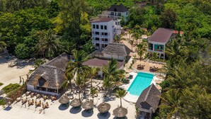 An aerial view of a resort with multiple buildings, a swimming pool, and beach areas with thatched-roof structures and palm trees. - Marea Boutique Hotel (Jambiani)
