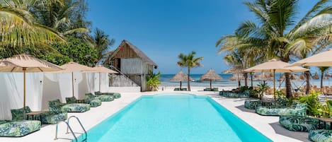 Ein Poolbereich mit Liegestühlen und Sonnenschirmen, umgeben von Palmen und Blick auf den Strand.