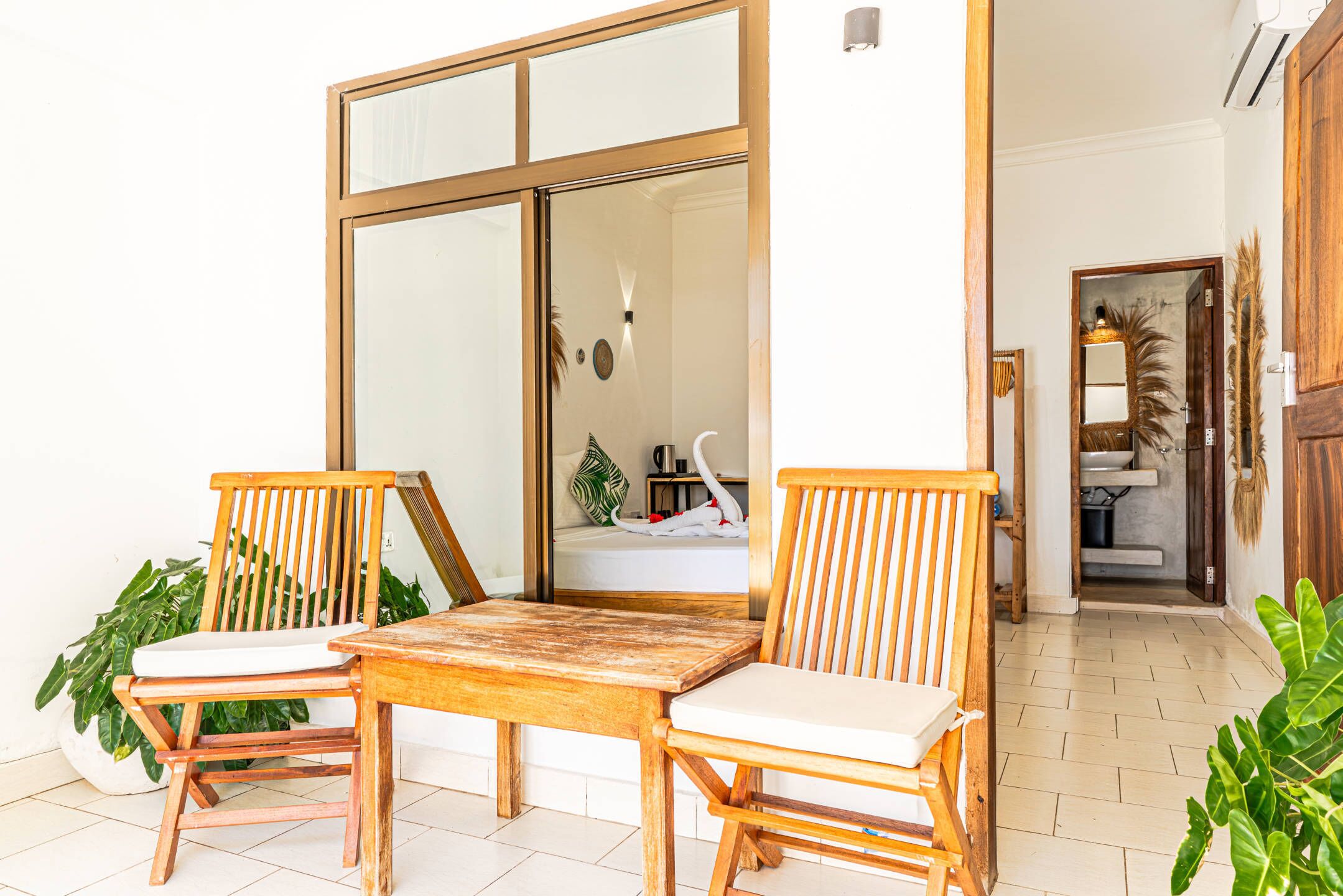 A wooden table and two chairs in a room with a tiled floor, a large sliding glass door, and a view of a bathroom with a bathtub and a mirror.