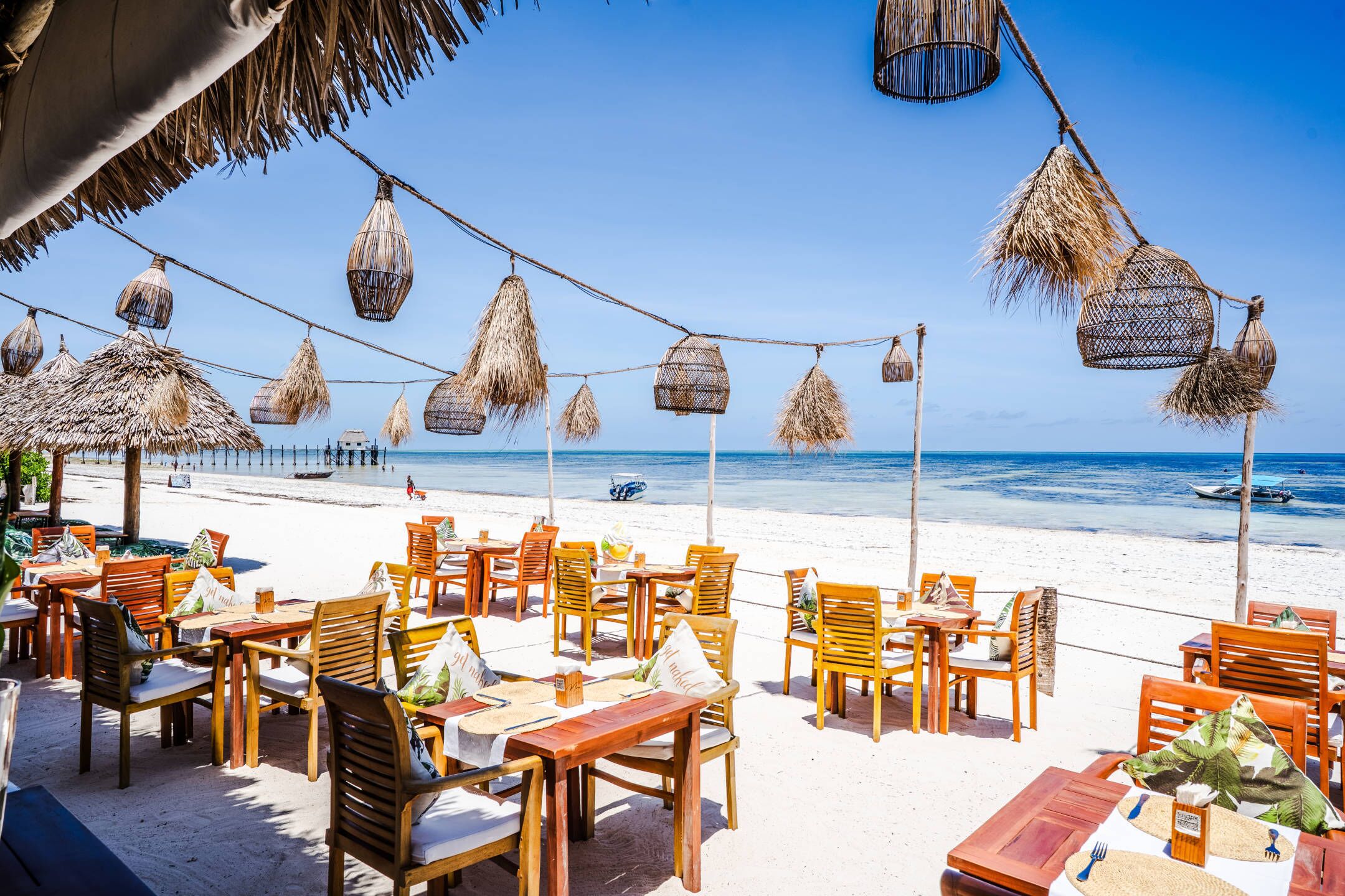 A beachside restaurant with wooden tables and chairs, thatched umbrellas, and hanging lanterns.