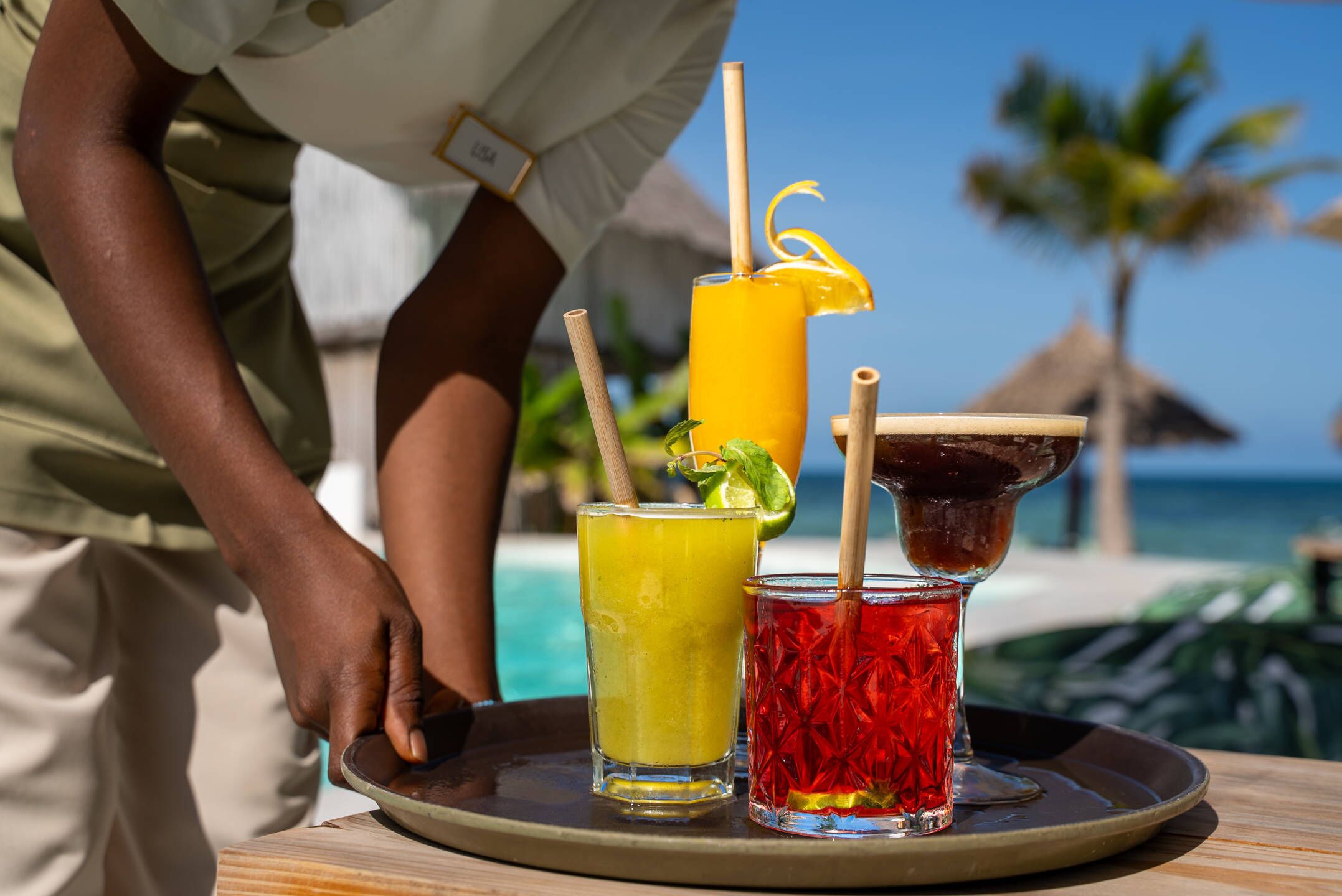 Una bandeja con tres bebidas: un cóctel amarillo, un cóctel rojo y una bebida oscura adornada con lima, sobre una mesa de madera junto a una piscina, con palmeras y un cielo despejado al fondo.