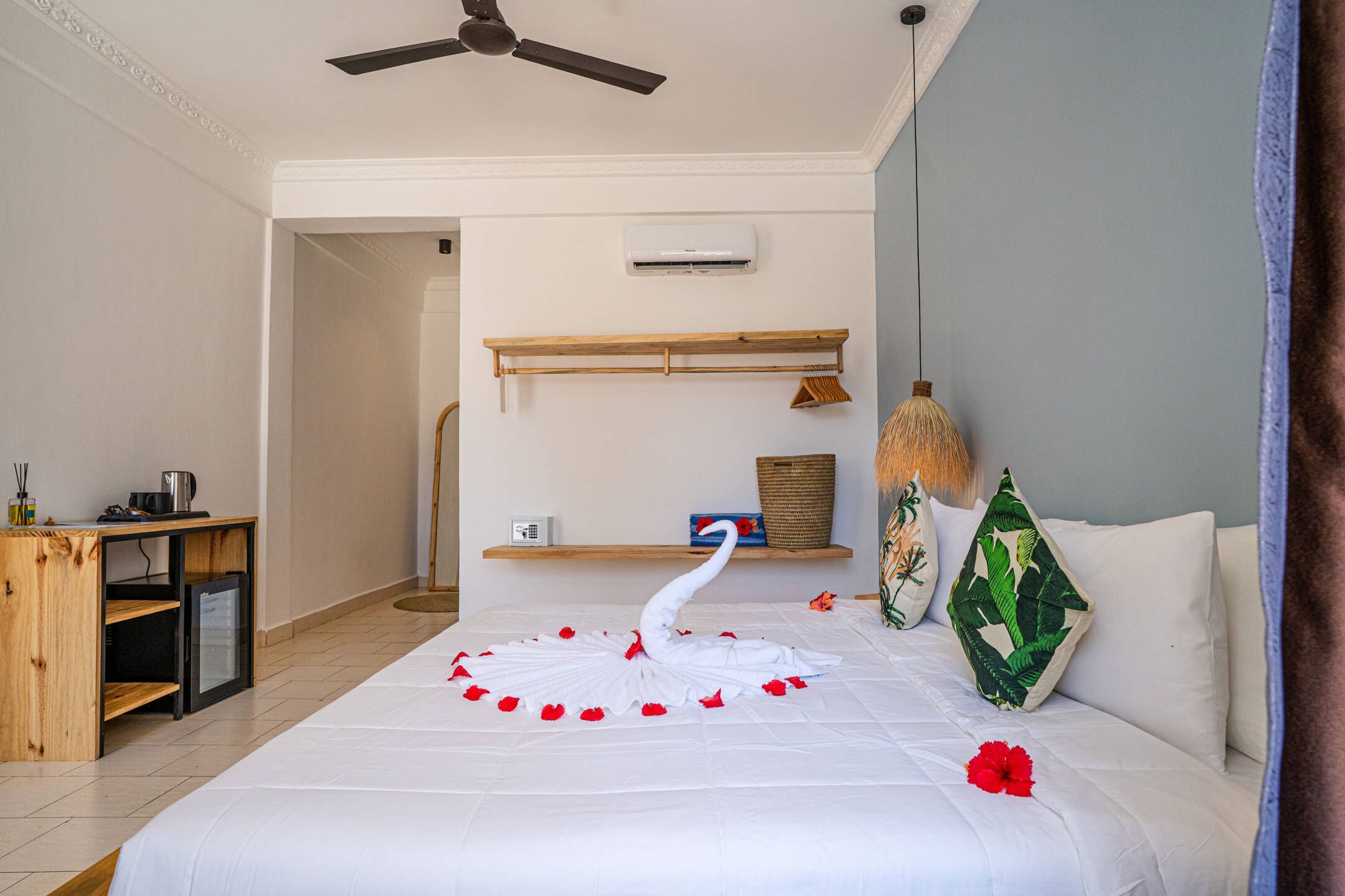A hotel room with a bed, a ceiling fan, a small table with a lamp, and a shelf with decorative items.