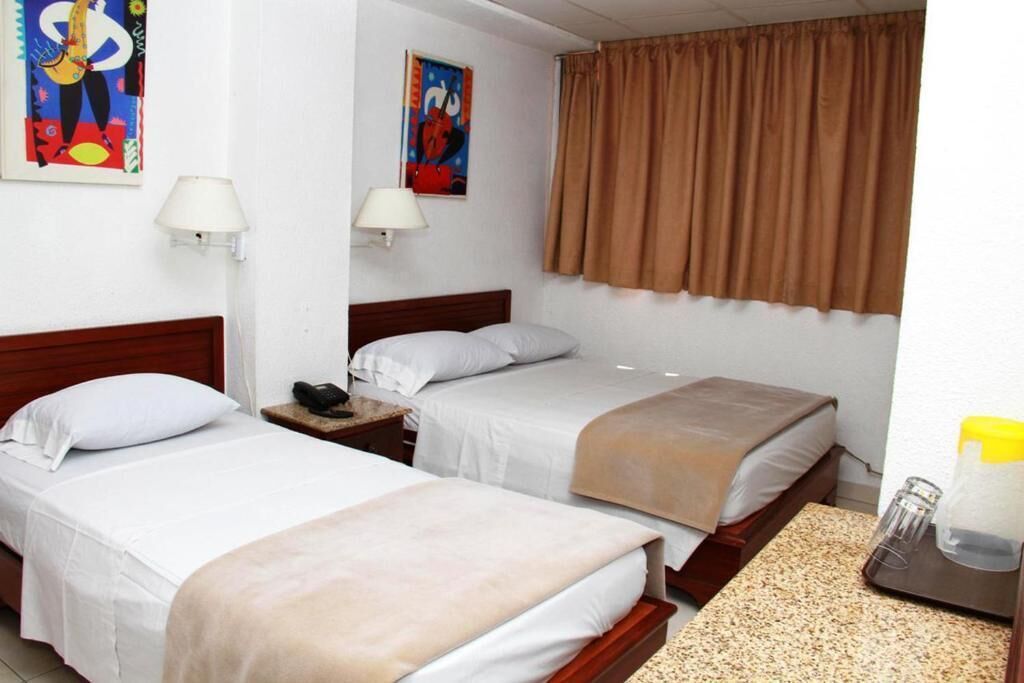 Ein Hotelzimmer mit zwei Einzelbetten, einem hölzernen Kopfteil, einem Nachttisch mit Lampe und einem Fenster mit braunen Vorhängen.