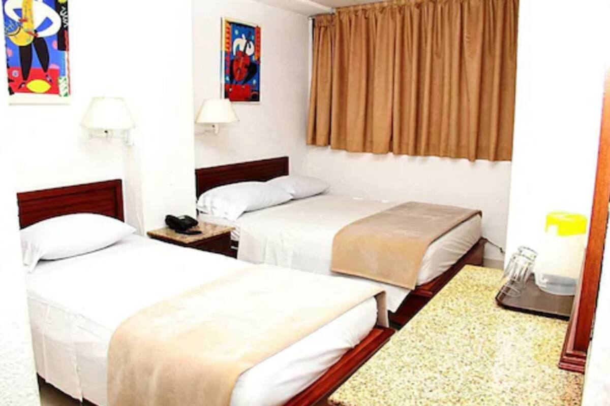 Une chambre d’hôtel avec deux lits simples, une tête de lit en bois, une table de chevet avec une lampe, une fenêtre avec des rideaux marron et une peinture abstraite colorée au mur.