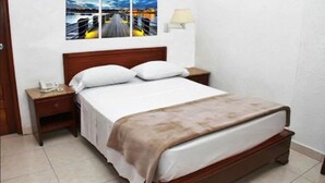 Una cama bien hecha con cabecera de madera, una mesita de noche con lámpara y un cuadro colgado en la pared que muestra un paisaje urbano.