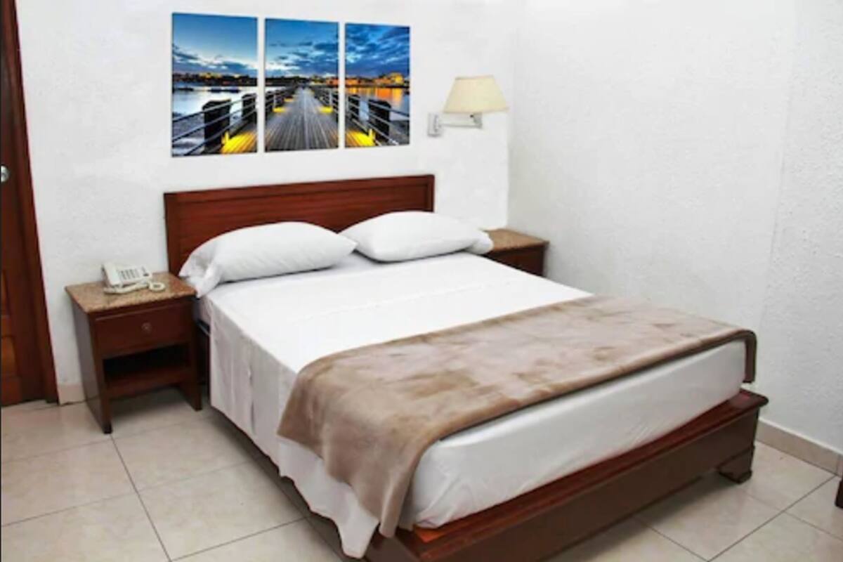 Un lit bien fait, avec une tête de lit en bois, une table de chevet avec une lampe et un tableau représentant un paysage urbain accroché au mur.