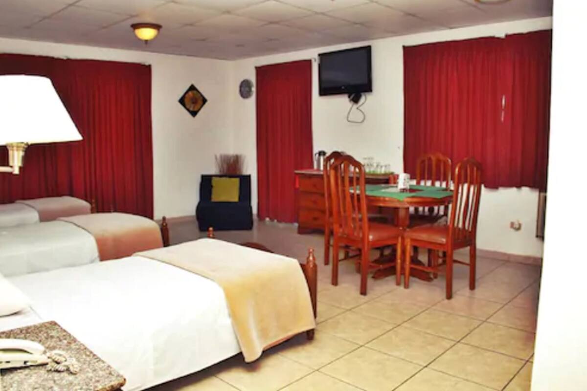 Une chambre d’hôtel avec deux lits, une table à manger avec des chaises, un téléviseur et des rideaux rouges.