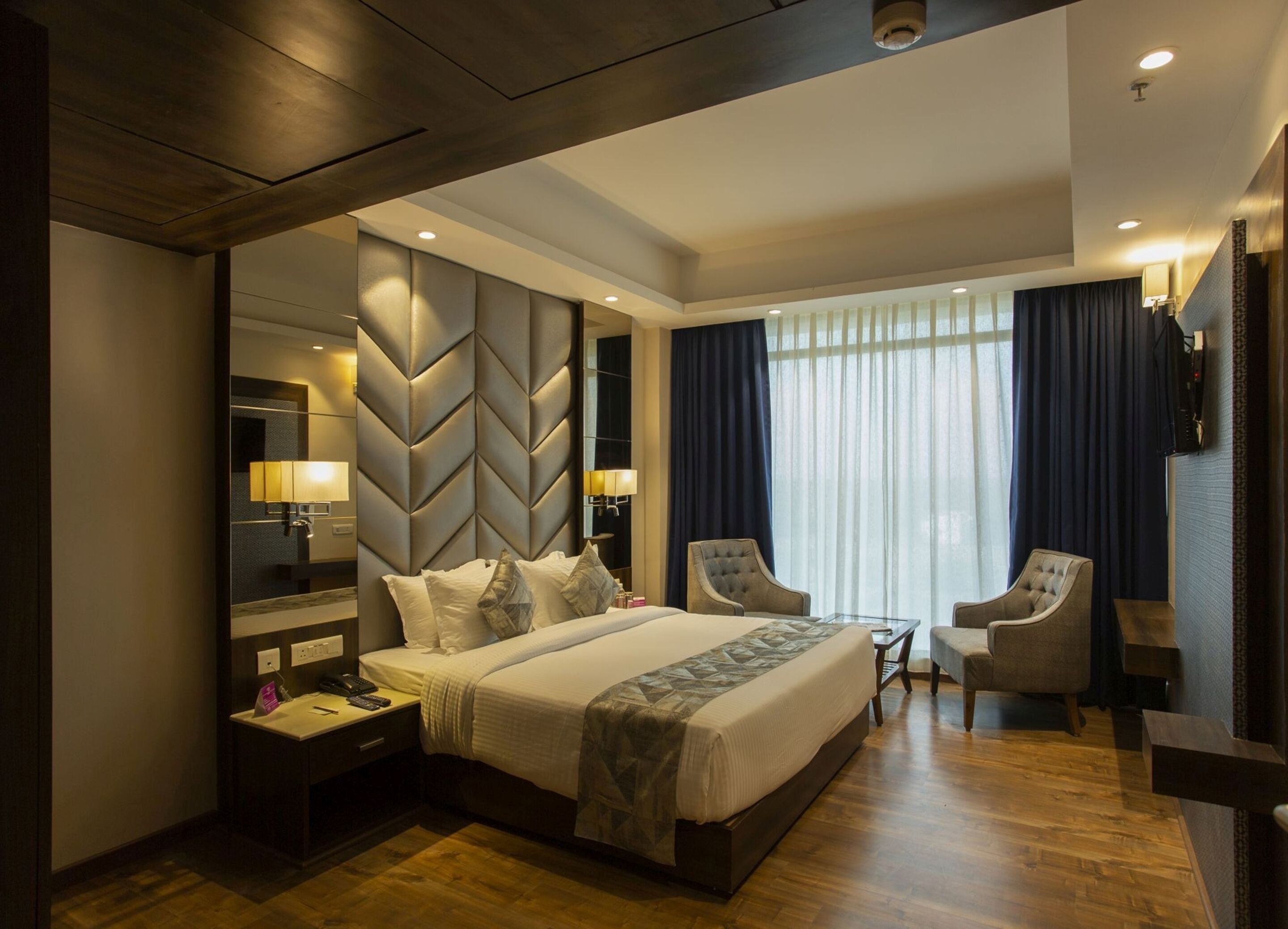 A modern hotel room with a large bed, two armchairs, a small table, and a large mirror.