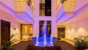 A hotel lobby with a central fountain, a reception area, and a staircase. - Sarovar Portico Somnath (Veraval)